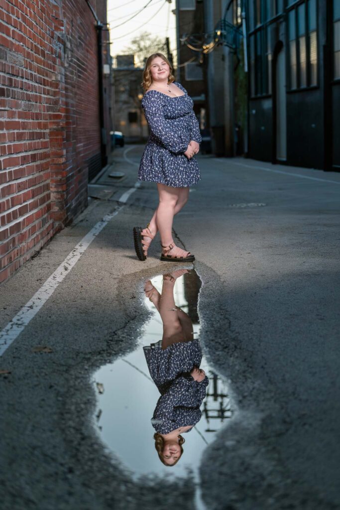 High school senior girl standing in urban ally with reflection in puddle on street.